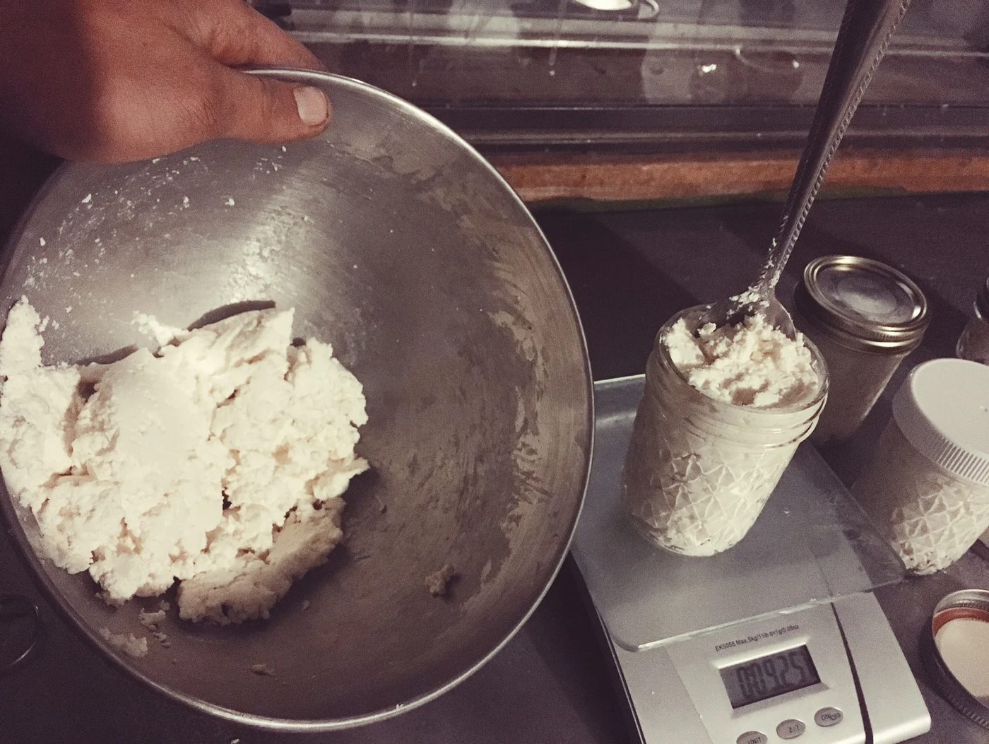 A hand tipping a steel bowl of fresh chevre beside a jar being filled on a kitchen scale
